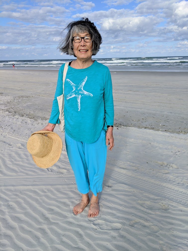 Yukiko Shuya smiling on the beach in a turquoise starfish shirt, holding a straw sun hat